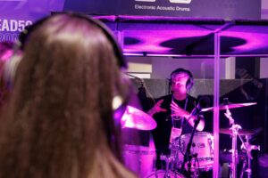A group of people gathered around a man in a glass booth wearing headphones and playing a set of drums.