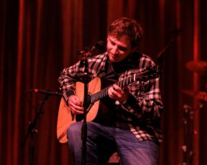 A seated man in a flannel plaid shirt playing an acoustic guitar.