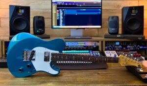 A blue single cutaway electric guitar on its side in front of a mixing desk, a computer monitor, and four speakers.