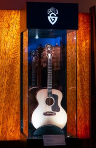 A Guild acoustic guitar on display behind glass at a trade show.
