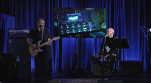 Two men on a stage in front of a screen at a trade show, one playing guitar, one talking into a microphone.