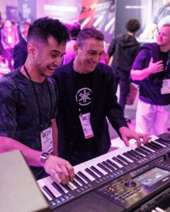 Two men at an electronic keyboard in a trade show, both smiling.