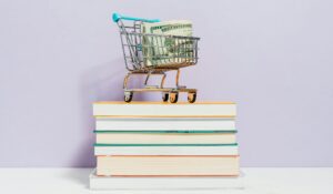 pile of books with a small shopping cart on top filled with money 