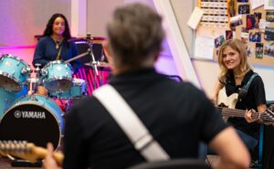 music educator in foreground facing students playing guitar and drum set