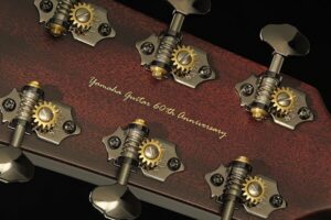 The back of the headstock of an acoustic guitar showing gold tuners and a gold inscription reading "Yamaha Guitars 60th Anniversary"